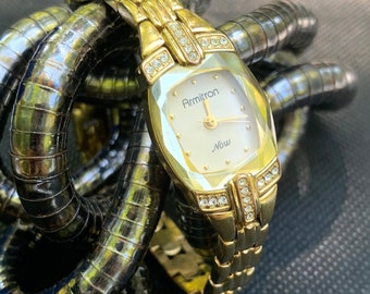 Può includere: Un orologio Armitron dorato con un quadrante bianco e accenti di cristallo. Il quadrante dell'orologio ha le parole "Armitron" e "Now" stampate su di esso.