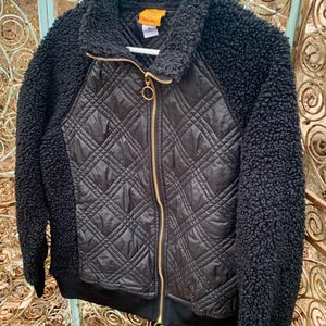May include: Black jacket with a quilted front panel and a plush, textured collar and sleeves. The jacket features a gold-toned zipper and a ribbed hem. The jacket is hanging on a white hanger.