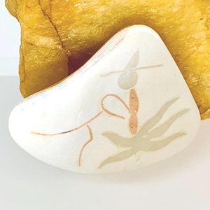 Puede incluir: Broche blanco de forma triangular con un diseño pintado. El diseño presenta una figura con sombrero, una línea marrón claro y una forma floral beige claro. La broche se encuentra sobre un fondo amarillo.