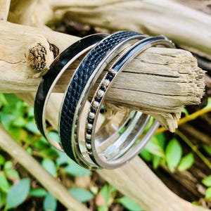 May include: A collection of silver bangles with black accents, including a textured band and a band with small silver beads. The bracelets are displayed on a piece of driftwood against a backdrop of green foliage.