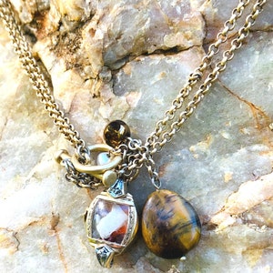 May include: A gold chain necklace with a charm featuring a clear crystal with brown and white inclusions, a brown tiger's eye stone, and a gold clasp.
