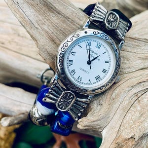 May include: A silver wristwatch with a white face and Roman numeral hour markers. The watch has a silver butterfly design on the band and a blue and white beaded strap.