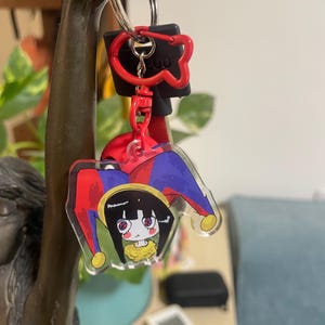 May include: A clear acrylic keychain featuring a cartoon character with a jester hat in red, blue, and yellow. The keychain is attached to a red clasp and a black key holder. The background includes a bronze sculpture and a green plant.