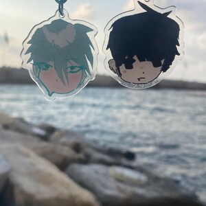 Op de afbeelding: Twee doorzichtige acryl sleutelhangers met geïllustreerde anime-stijl karakterhoofden. De linker sleutelhanger toont een personage met lichtgroen haar en roze wangen, de rechter sleutelhanger toont een personage met zwart haar en een neutrale blik.