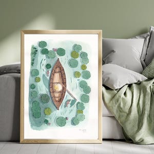 Op de afbeelding: Aquarel schilderij van een houten roeiboot op een vijver, omringd door waterlelies. De boot is bruin met een gele cirkel in het midden. Het water is lichtblauw en de waterlelies zijn groen. Het kunstwerk is ingelijst in een licht houten frame.