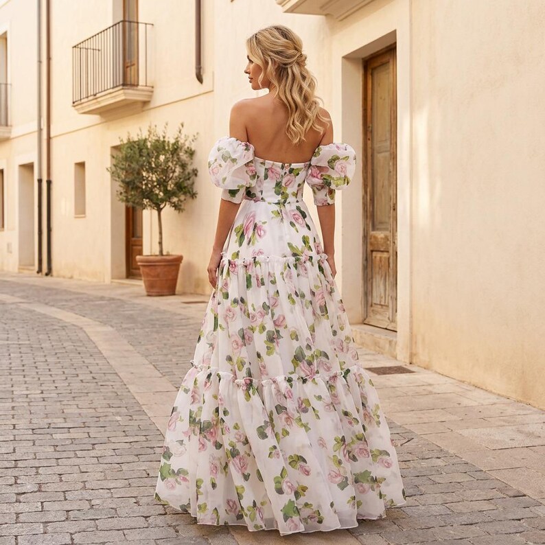 May include: A white, off-the-shoulder gown with a floral pattern of pink and green roses. The dress has puff sleeves and a tiered skirt, creating a flowing silhouette. The dress is worn outdoors in a cobbled street.
