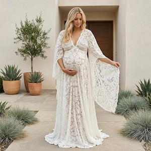 May include: A white lace maternity dress with a deep V-neck and flowing sleeves. The dress has detailed floral patterns and a floor-length skirt. The expectant parent is holding their belly. The background includes plants and a building.