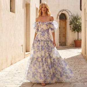May include: A cream-coloured, off-the-shoulder, floor-length gown with a purple floral pattern. The dress has short, puffy sleeves and a tiered skirt. Suitable for special occasions, the dress is a beautiful example of formal wear.