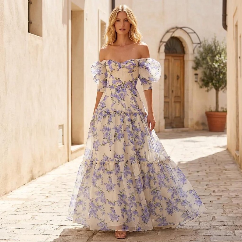 May include: A cream-coloured, off-the-shoulder, floor-length gown with a purple floral pattern. The dress has short, puffy sleeves and a tiered skirt. Suitable for special occasions, the dress is a beautiful example of formal wear.