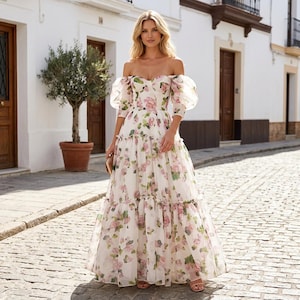 May include: A white, off-the-shoulder maxi dress adorned with a floral print of pink and green roses. The dress has puff sleeves and a tiered skirt, creating a romantic and elegant style. Suitable for a special occasion.