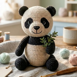 May include: A handmade crochet panda bear with black and cream yarn. The panda has black ears, eyes, and limbs. It holds a sprig of greenery. The bear sits on a gray linen cloth with yarn balls, a crochet hook, and a wooden tag.