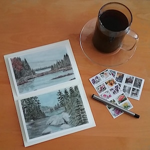 May include: A still-life image featuring two landscape watercolor paintings, a clear glass mug of coffee on a saucer, and several decorative stamps. A silver pen rests near the stamps. The paintings depict serene forest and water scenes.
