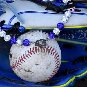 May include: A baseball with red stitching and a silver charm with the number 43 is displayed on a blue and white baseball glove. A necklace with purple and clear crystal beads is draped over the baseball and glove.