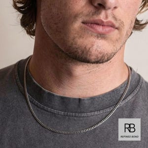 Könnte beinhalten: Eine silberne Halskette mit Curb-Kettengliedern. Die Halskette wird von einer Person getragen, die ein graues T-Shirt trägt. Das Bild enthält das Logo von Refined Bond.