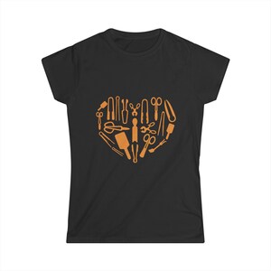 Puede incluir: Camiseta negra con un diseño en forma de corazón compuesto por herramientas de color naranja, como tijeras y peines. La camiseta está hecha de un material suave. Las herramientas están dispuestas en forma de corazón.