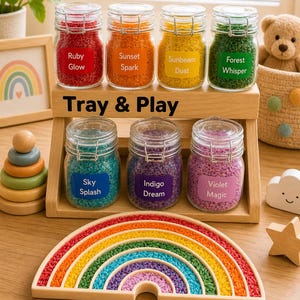 May include: A wooden tray holding seven glass jars filled with coloured sensory materials. The jars are labelled "Ruby Glow," "Sunset Spark," "Sunbeam Dust," "Forest Whisper," "Sky Splash," "Indigo Dream," and "Violet Magic." A rainbow-shaped wooden toy is in the foreground.