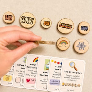 May include: A set of circular wooden discs featuring weather-themed illustrations and text including RAIN, CLOUDY, SUN, RAINBOW, and SNOW, accompanied by a series of instructional cards with prompts like FIND ALL THE DISCS and WHAT WEATHER.