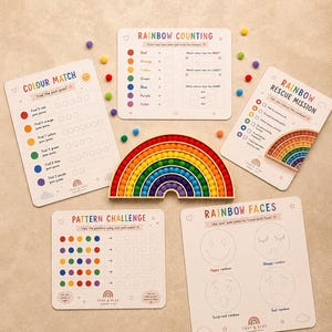 May include: Educational toys including a wooden rainbow puzzle and activity cards. The cards offer colour matching, counting, pattern challenges, and rainbow face tracing. Scattered around are colourful pom-poms.