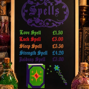 May include: A wooden-framed chalkboard displaying the word "Spells" in purple script. The board lists spells with prices in pounds: Love, Luck, Sleep, Strength, and Jaidens. An illustration of a book and wand are included.