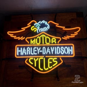 May include: A neon sign featuring the Harley-Davidson logo with an eagle emblem at the top. The sign displays the text MOTOR HARLEY-DAVIDSON CYCLES in bright yellow, white, and orange neon tubing against a dark background, creating a glowing effect.