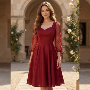 May include: A knee-length burgundy dress featuring a sweetheart neckline and sheer long sleeves with a polka dot pattern. The sleeves are finished with delicate tie-up cuffs, and the skirt has a full, flared silhouette that falls from the waist.