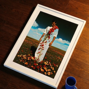 Könnte beinhalten: Ein gerahmter Kunstdruck mit einer Person in Weiß, geschmückt mit roten Blumen, vor einem Feld mit Wildblumen unter blauem Himmel. Eine blaue Tasse steht daneben auf einer dunklen Holzoberfläche.