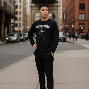 Op de afbeelding: Zwart sweatshirt met lange mouwen met de tekst "FISTS OF FURY" en een witte afbeelding van een persoon in een vechthouding. De persoon draagt ook een zwarte broek en witte sneakers. De achtergrond is een stadsstraat.