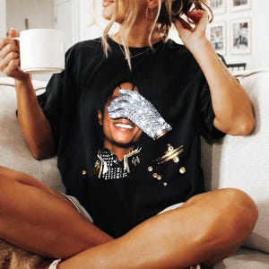 May include: Black t-shirt featuring a graphic of Michael Jackson with a sparkling glove covering his eyes. The shirt also has gold and rhinestone embellishments. The person in the image is holding a white mug.
