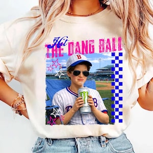 Puede incluir: Camiseta color crema con el texto "Hit the Dang Ball" en rosa y azul. La camiseta presenta un gráfico de una persona con uniforme de béisbol sosteniendo una bebida Monster Energy. La persona lleva gafas de sol y una gorra de béisbol.
