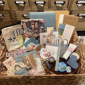 May include: A woven basket filled with reading accessories including a floral notebook, a canvas pouch with text, a reading list card, sticky notes, bookmarks, a hand cream tube, a shower steamer, a small blue bag charm, and various stickers.