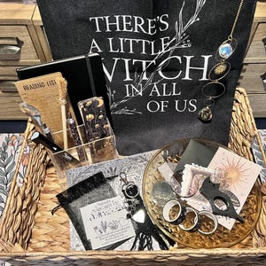 May include: A woven basket containing a black tote bag with the text THERE'S A LITTLE WITCH IN ALL OF US, a black notebook, a reading list card, a clear acrylic organizer, a sun and moon suncatcher, a glass dish, washi tape, and stationery.