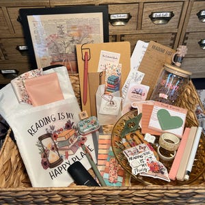 May include: A wicker basket filled with reading-themed items. Includes a tote bag with the text "Reading is my happy place", bookmarks, pens, stickers, a glass jar, and stationery. The items are in shades of pink, peach, and green.