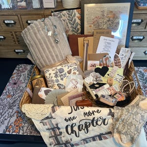 May include: A wicker basket filled with reading-themed items, including a soft, gray blanket, journals, decorative cards, and a candle. A floral patterned mat and socks are also visible. The text "just one more chapter" is printed on a white cloth.
