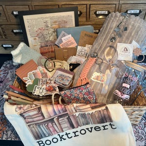 May include: A wicker basket overflowing with book-themed gifts. Includes a tote bag with a bookshelf graphic, pens, stickers, a jar, and a framed print. The basket sits on a floral patterned mat.