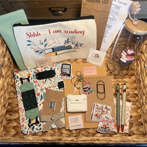 May include: A woven basket filled with reading-themed items. Includes a canvas pouch with "Shhh... I am reading" text, a glass tumbler, pens, stickers, a bookmark, and a reading list. The items are arranged on a floral patterned surface.