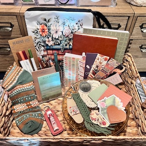 May include: A woven basket containing a floral print pouch, a brown notebook, a green grid notebook, a reading list card, colored markers, a book review card, patterned bookmarks, a green patterned sock, lip balm, and a green macrame keychain.