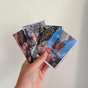 May include: A hand holding a fan of tarot zines. The zines have various colorful collage designs and text, including "SWORDS" and "A TAROT ZINE". The background is a plain white wall.