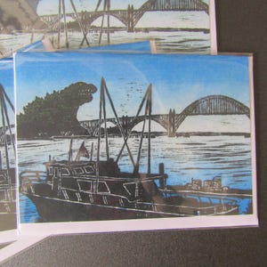 May include: A rectangular greeting card featuring a black ink block print on a blue background. The illustration depicts a large sea creature rising behind a bridge and a boat in the foreground. The scene is enclosed in a clear protective sleeve.