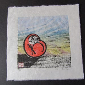 May include: A Daruma doll print on textured paper. The red doll has black details, set against a blue and yellow landscape. The print is signed and numbered 14/50. The paper has a textured, handmade appearance.