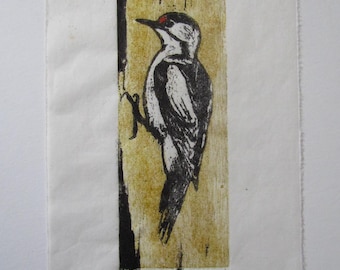 May include: A woodblock print showing a woodpecker on a tree trunk. The bird is in black and white with a red head spot. The background is light brown, and the print is on white paper. Signed by the artist.