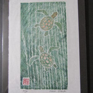 May include: A framed art print depicting two sea turtles against a green, textured backdrop. The turtles have green flippers and patterned shells with orange and pink details. Signed by Keori Clark, with a small red stamp and the number 14/80.