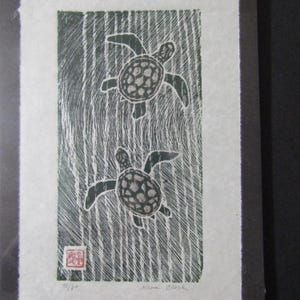May include: A framed print depicting two sea turtles against a textured, vertical background. The turtles are dark with patterned shells. The print includes a red stamp, the text "11/80" and the artist's signature "Kira Clark".