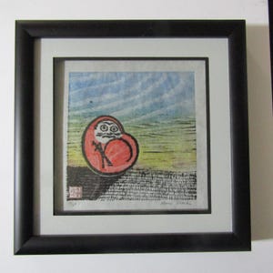 May include: Framed print of a Daruma doll, a traditional Japanese good luck charm. The doll is red with a white face and black eyes, set against a blue and green background. The print is in a black frame.