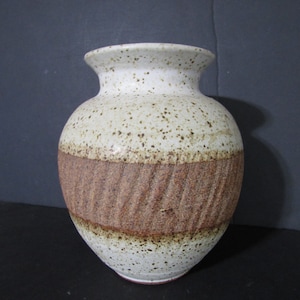 May include: A white ceramic vase with brown speckles and a brown band around the middle. The vase has a rounded body and a slightly flared opening.
