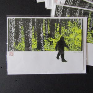 May include: A black and white art print of a Bigfoot walking in a forest. The background features tall, black trees and bright green foliage. The foreground is white, with the silhouette of Bigfoot. The print is on a white card.