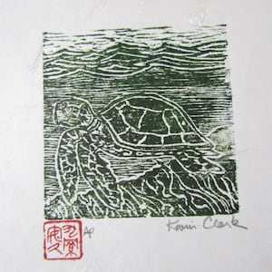 May include: A green and white linocut print depicting a sea turtle in the ocean. The print is signed "Kevin Clark" and features a red chop mark. The turtle's shell pattern is visible, set against stylized waves. The artwork is on white paper.