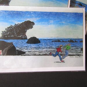 May include: A rectangular greeting card featuring a colorful illustration of two children running along a beach. In the background, a large, dark, jagged rock formation resembling a monster rises from the blue ocean under a clear sky.