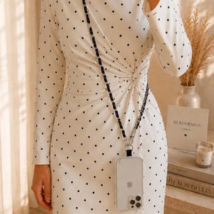 May include: A white long-sleeved dress featuring a black polka dot pattern and a side-gathered knot detail at the waist. A clear smartphone case with a black and silver chain strap is worn across the body over the dress.