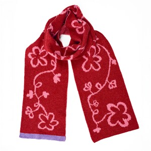 May include: A red knitted scarf with a pink floral design. The scarf features a purple border at the ends. The pattern includes stylized flowers and vines. The scarf is made of a soft, textured material.