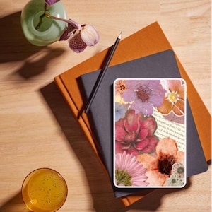 Puede incluir: Una composición floral con una funda para tableta con un collage de flores en tonos rosas, naranjas y morados. También hay un jarrón con orquídeas moradas, un lápiz, un cuaderno y un vaso de líquido naranja.